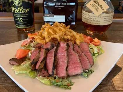 Sliced steak salad with onion straws in front of Kentucky bourbon bottles